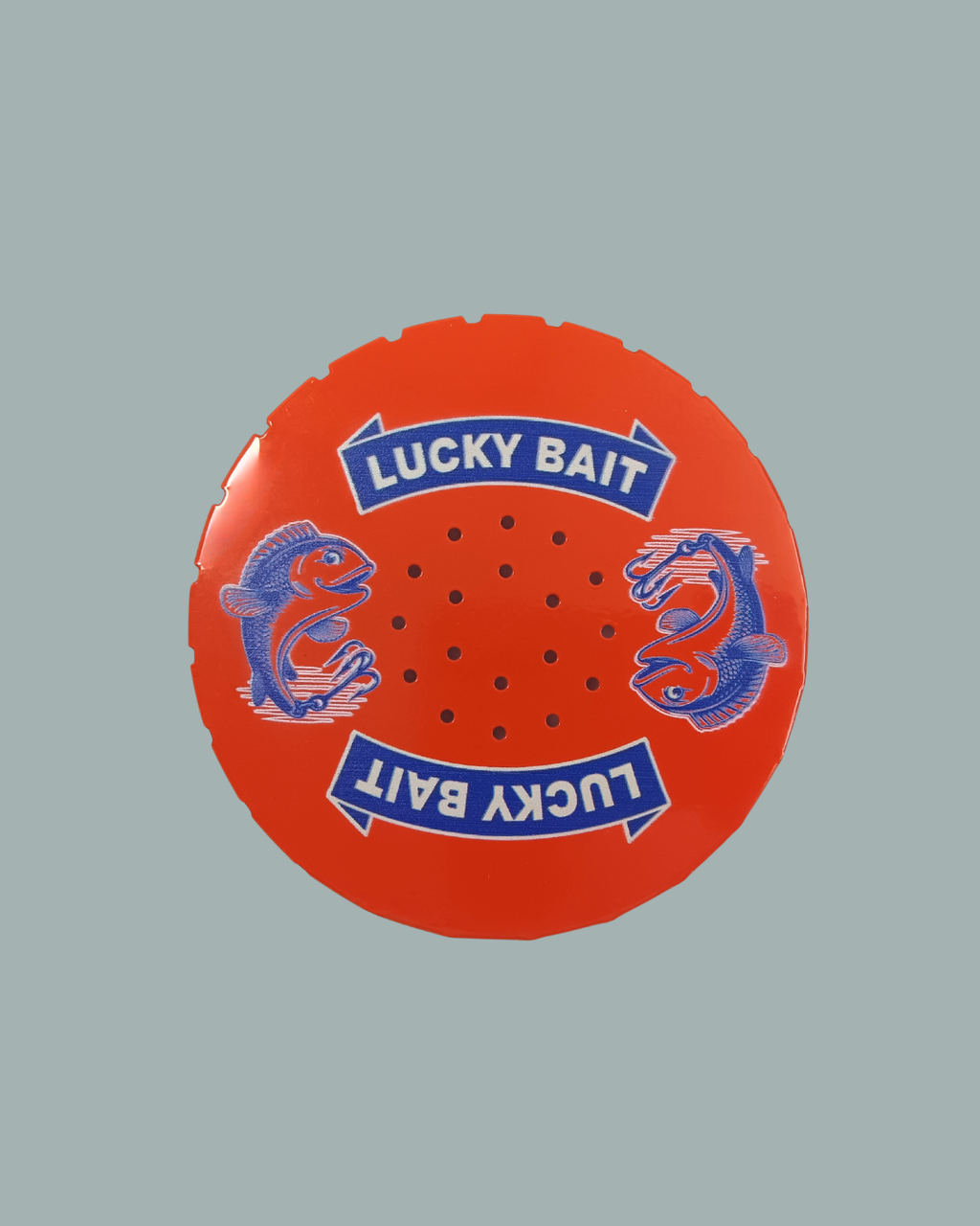 Lucky Bait Betesburk från Lucky Bait Shop har ett runt rött lock med texten ”LUCKY BAIT” tryckt två gånger i vitt samt blå fiskillustrationer, flera små hål i mitten och avbildas mot en enfärgad grå bakgrund.