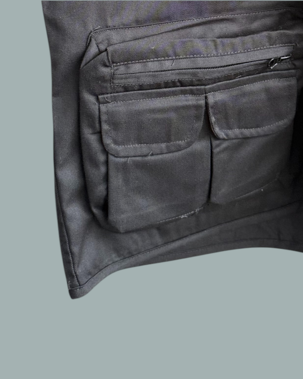 Närbild på sidan av den svarta Fiskevästen, med två fickor  och en ficka med dragkedja. Visas mot en stålblå bakgrund.