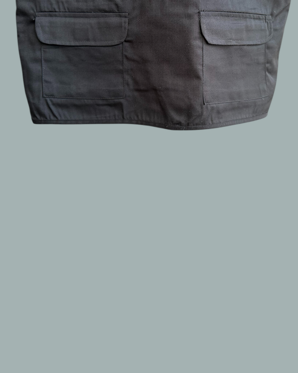 En närbild av den nedre delen av den svarta Fiskevästen, med två stora framfickor. Visas mot en stålblå bakgrund.