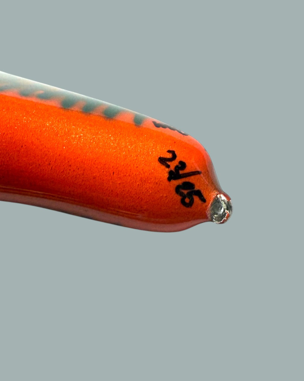 Närbild på spetsen på ett ljusorange Lucky Bait Limited Edition bete-fiskedrag, med svart text numrering, en blank yta och en liten genomskinlig öljett i änden. Visas mot en stålblå bakgrund.