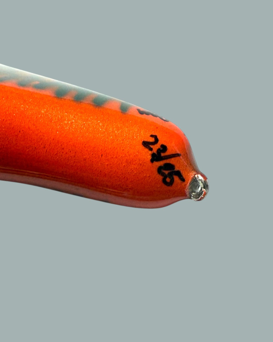 Närbild på spetsen på ett ljusorange Lucky Bait Limited Edition bete-fiskedrag, med svart text numrering, en blank yta och en liten genomskinlig öljett i änden. Visas mot en stålblå bakgrund.