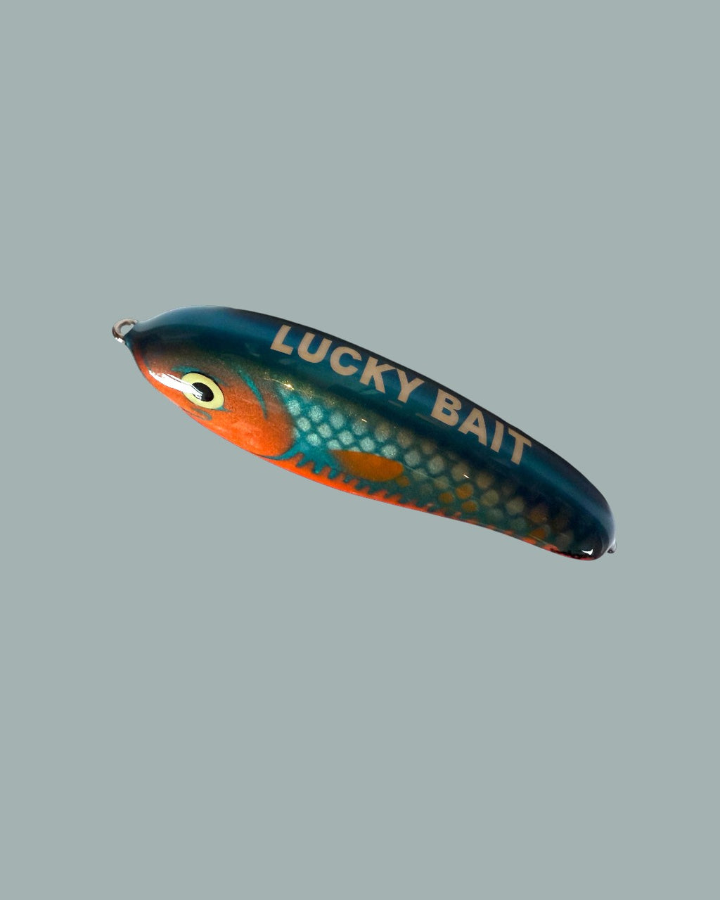 Lucky Bait Limited Edition-bete är ett målat fiskedrag med ett fiskfjällsmönster, ett illustrerat öga och "LUCKY BAIT" i fetstil med vita bokstäver på sidan. Visas mot en stålblå bakgrund.