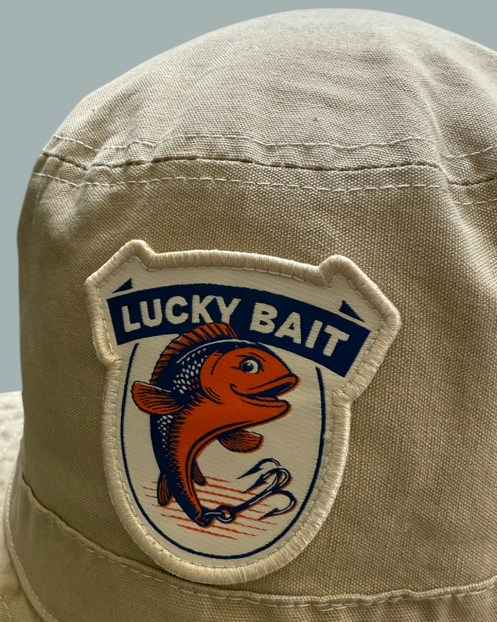 Närbild av den beigefärgade Fiskehatten från Lucky Bait, med en sköldformad patch med Lucky Bait logo, en orange fisk som hoppar över en krok.Visas mot en stålblå bakgrund.
