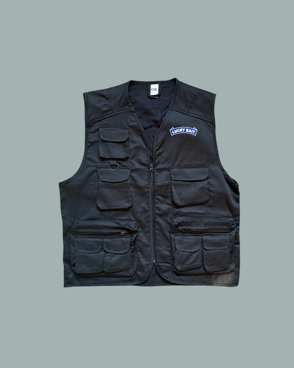Fiskevästen är en svart fiskeväst med flera fickor och en Lucky Bait-logo tryckt i vitt och blått på vänster bröstet, perfekt för din nästa fisketur. Visas mot en stålblå bakgrund.