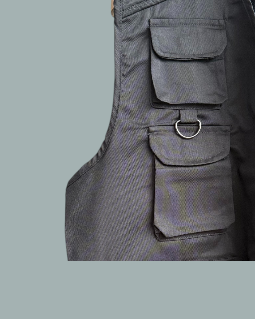 Närbild av Fiskeväst i svart, med flera fickor med lock och en D-ring i metall på framsidan. Visas mot en stålblå bakgrund.