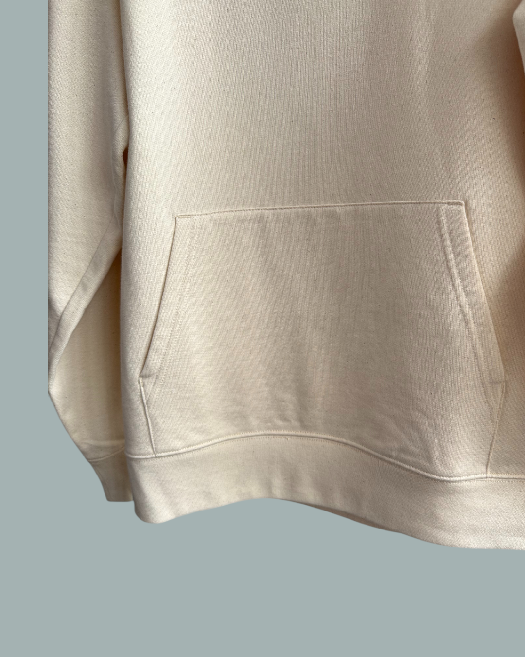 En närbild av Lucky Bait Hoodie Natur i beige med känguruficka fram. Visas mot en stålblå bakgrund.