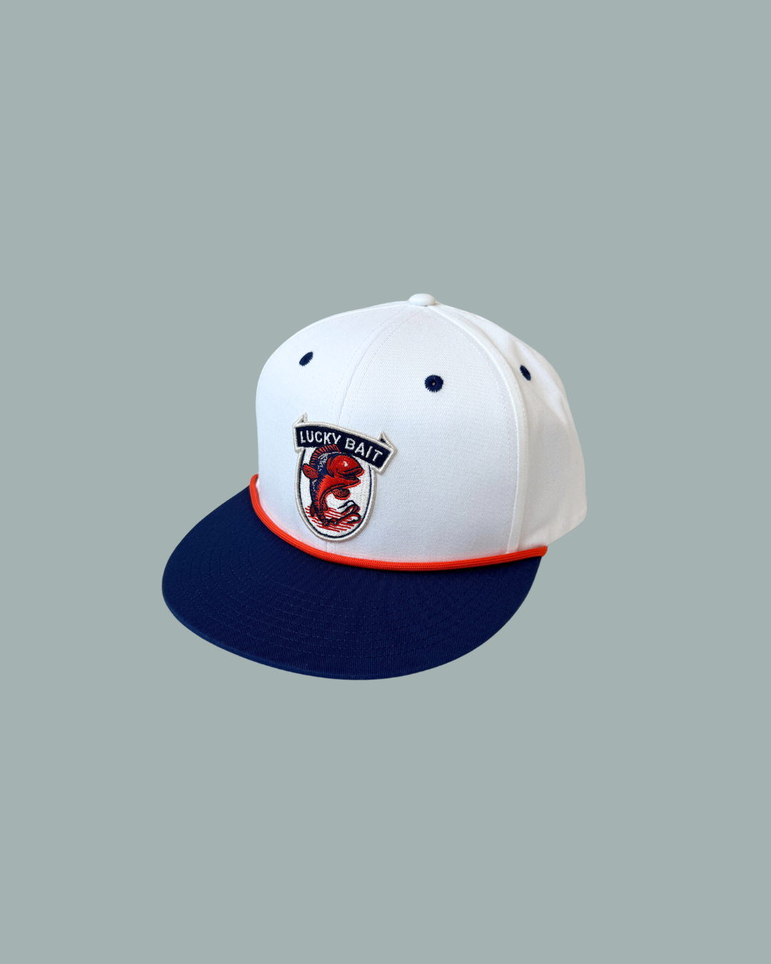 Lucky Bait Shops keps ”Lucky Bait Rope Cap” har en vit krona, marinblå skärm, orange kant och en logotyp i form av en fisk – perfekt för fisketurer – som framhävs mot en enfärgad grå bakgrund och ger en fräsch, modern look.