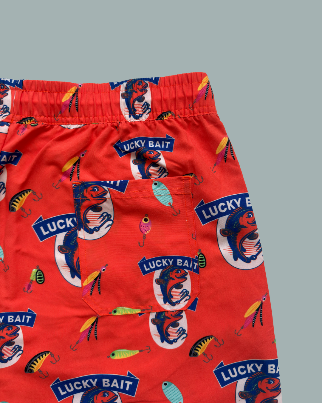 Lucky Bait Badbrallor från Lucky Bait Shop är färgglada badshorts med ficka, mönster med fiskedrag och logotyper med fiskmotiv, resår i midjan och visas här på grå bakgrund.