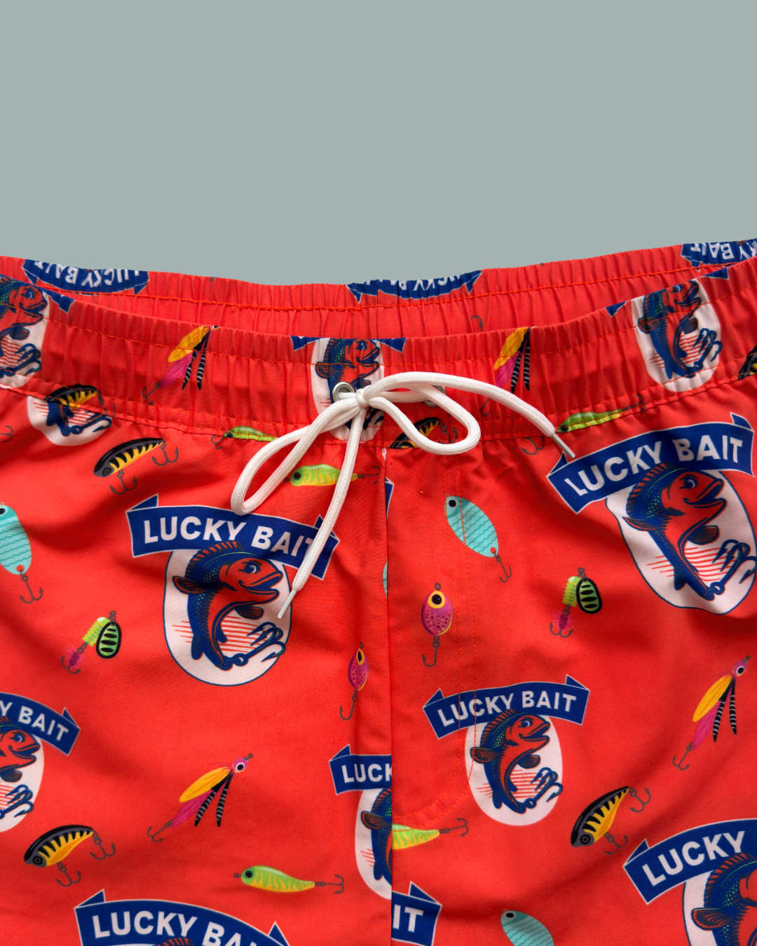 Lucky Bait Badbrallor från Lucky Bait Shop: röda badshorts med vit dragsko, färgglada mönster med fiskebeten och fisklogotyper – perfekta för dig som gillar färgglada badshorts. Visas mot en enfärgad bakgrund.