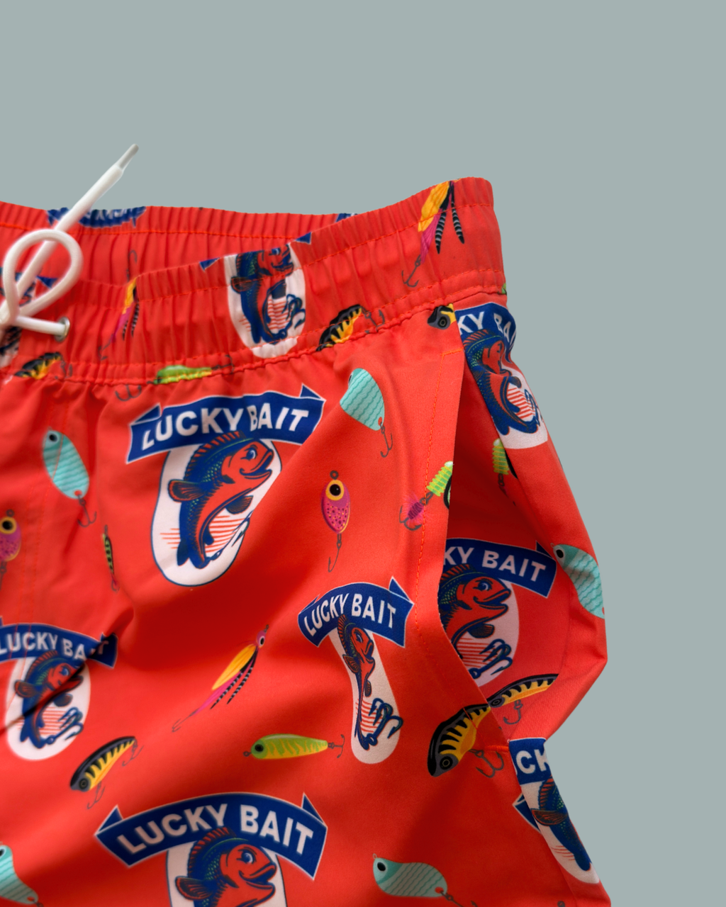 Lucky Bait Badbrallor från Lucky Bait Shop – färgglada badshorts för herrar med fisketema och livfulla fisk- och betesmönster samt texten ”Lucky Bait” på röd bakgrund, framhävda mot en enkel grå bakgrund.