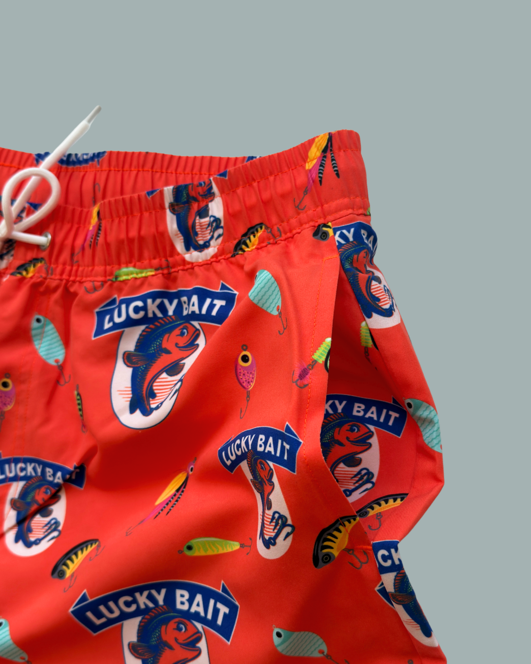 Lucky Bait Badbrallor från Lucky Bait Shop – färgglada badshorts för herrar med fisketema och livfulla fisk- och betesmönster samt texten ”Lucky Bait” på röd bakgrund, framhävda mot en enkel grå bakgrund.