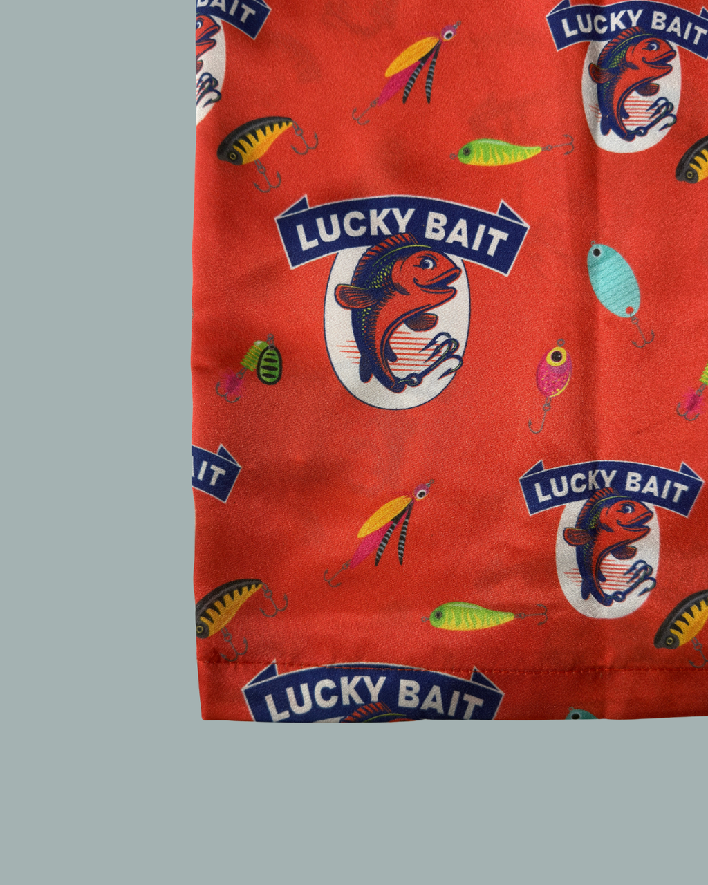 Skjorta NY från Lucky Bait är en röd tygskjorta med ett mönster av färgglada fiskebeten och logotyper som föreställer en fisk och texten ”Lucky Bait” på en vit oval bakgrund.