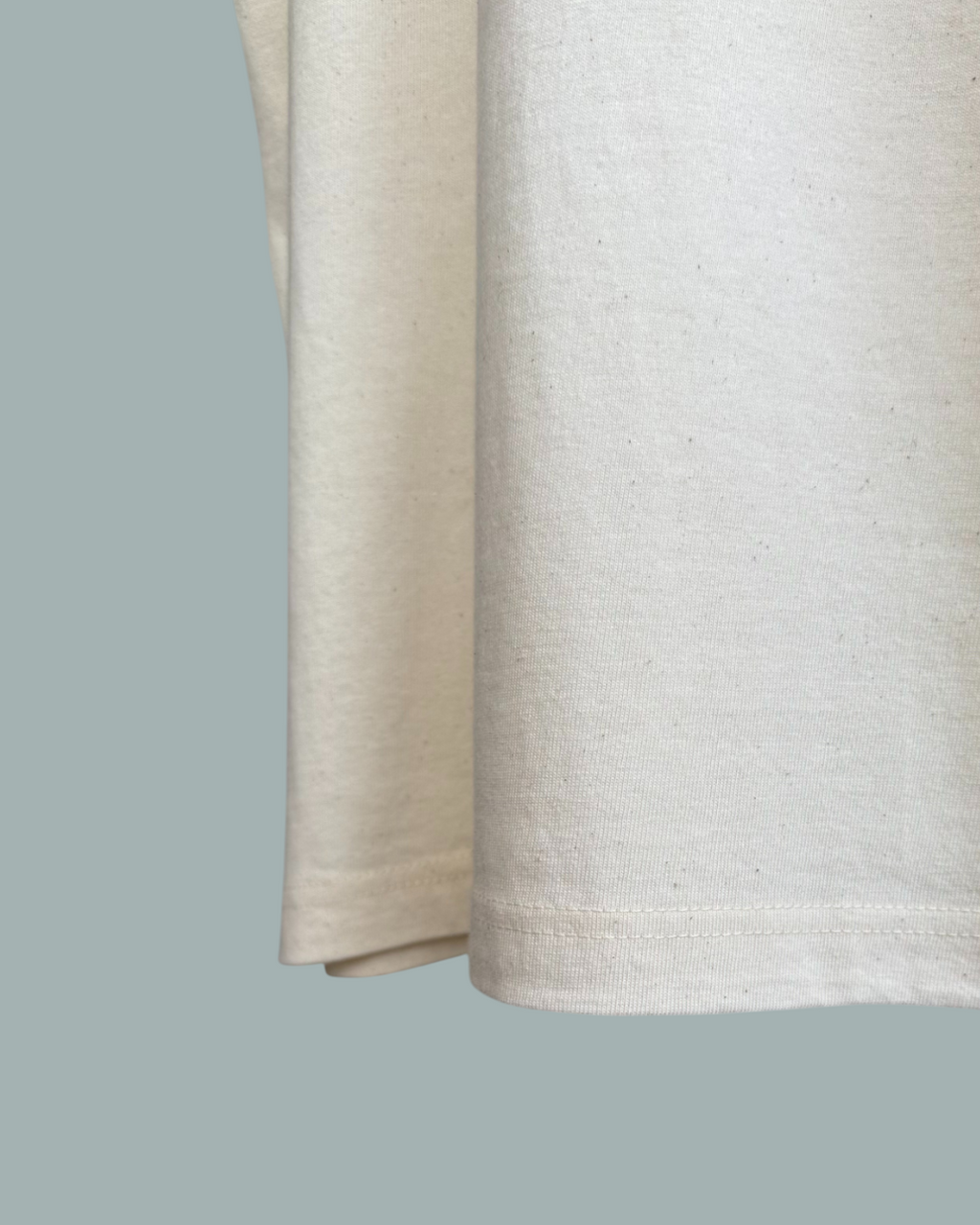 Närbild av fållen och undersidan på en Lucky Bait T-Shirt Natur i beige, med fokus på tygstrukturen och sömnadsdetaljerna. Endast en del av tröjan visas. Visas mot en stålblå bakgrund.