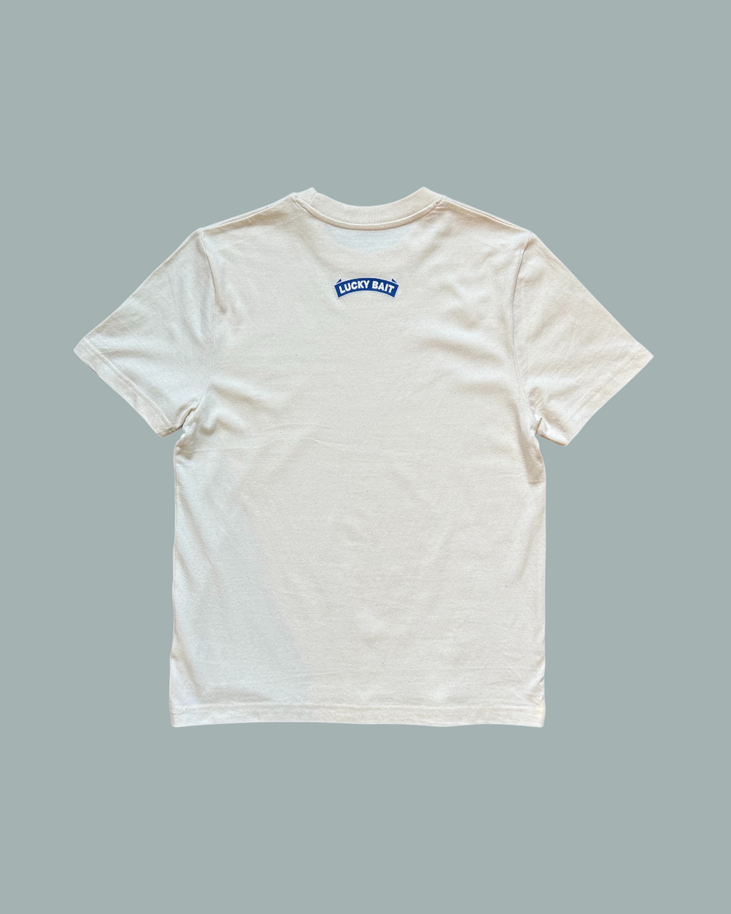 En enfärgad beige T-shirt Natur från Lucky Bait ligger platt med baksidan uppåt. Blåvit liten logo Lucky bait nedanför nacken. Visas mot en stålblå bakgrund..
