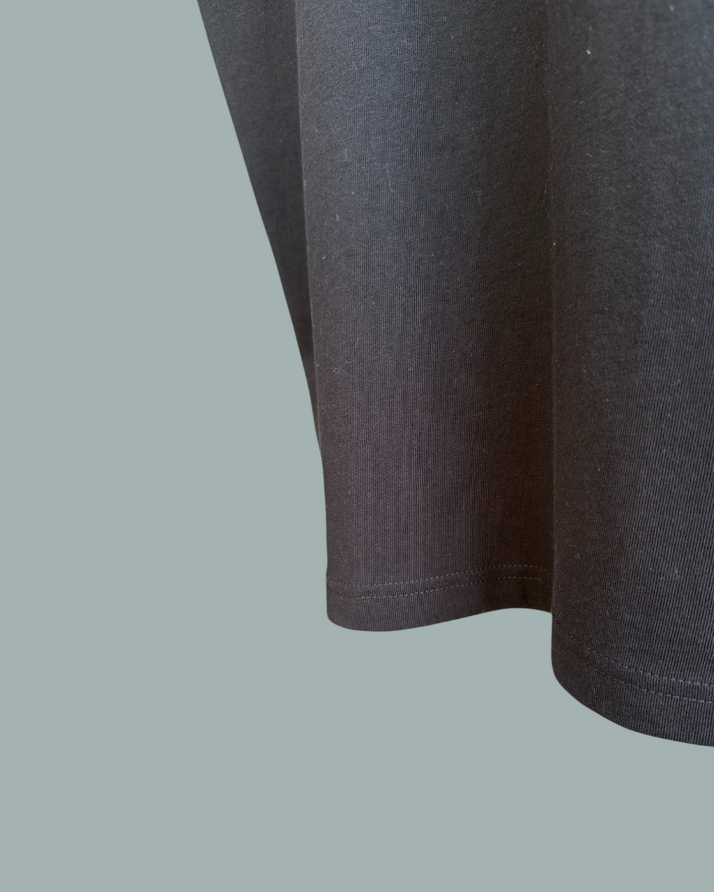 Närbild av fållen och undersidan på en Lucky Bait T-Shirt Svart, med fokus på tygstrukturen och sömnadsdetaljerna. Endast en del av tröjan visas. Visas mot en stålblå bakgrund.