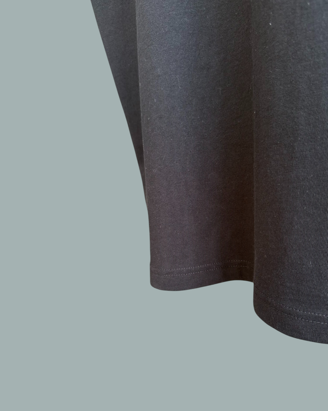 Närbild av fållen och undersidan på en Lucky Bait T-Shirt Svart, med fokus på tygstrukturen och sömnadsdetaljerna. Endast en del av tröjan visas. Visas mot en stålblå bakgrund.